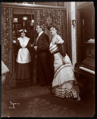 Un homme et une femme habillés formellement et ce qui semble être un domestique dans une pièce avec un piano, 1905 - Byron Company